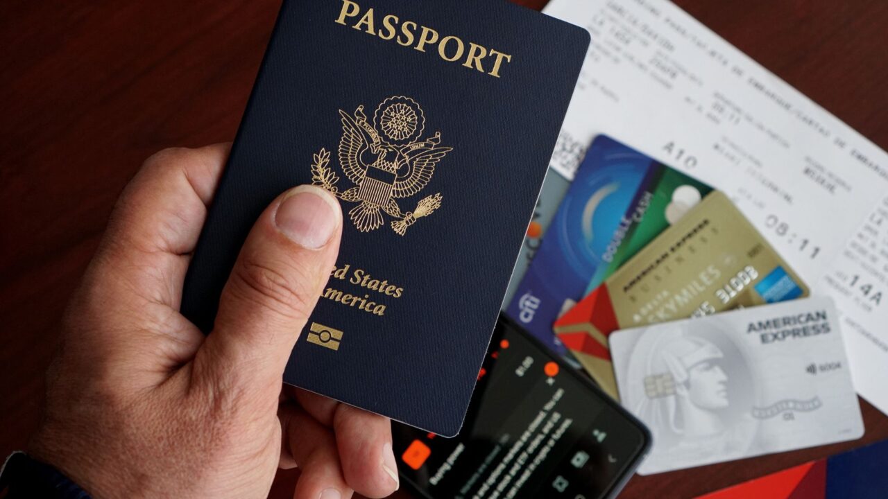 A passport held above a boarding pass, payment cards and a smartphone on a table