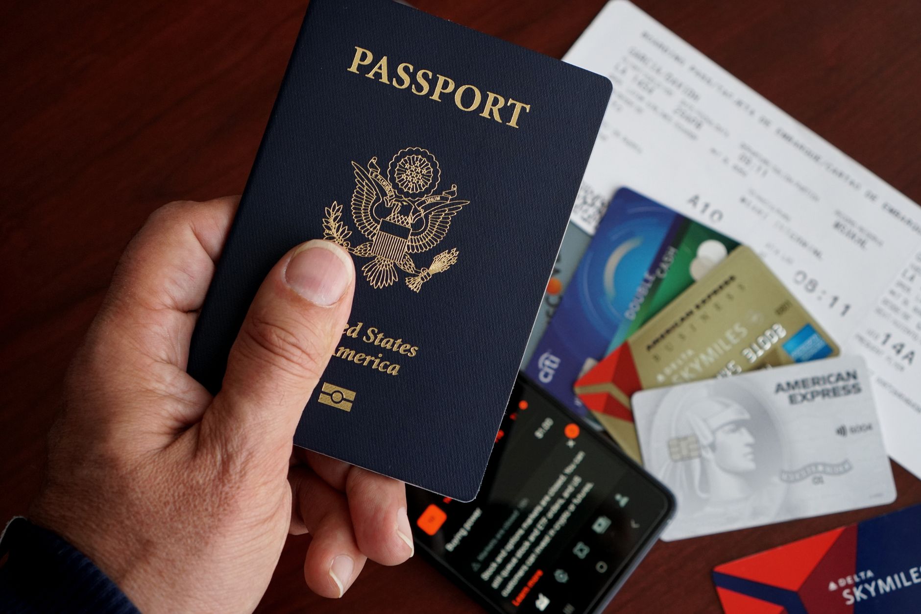 A passport held above a boarding pass, payment cards and a smartphone on a table