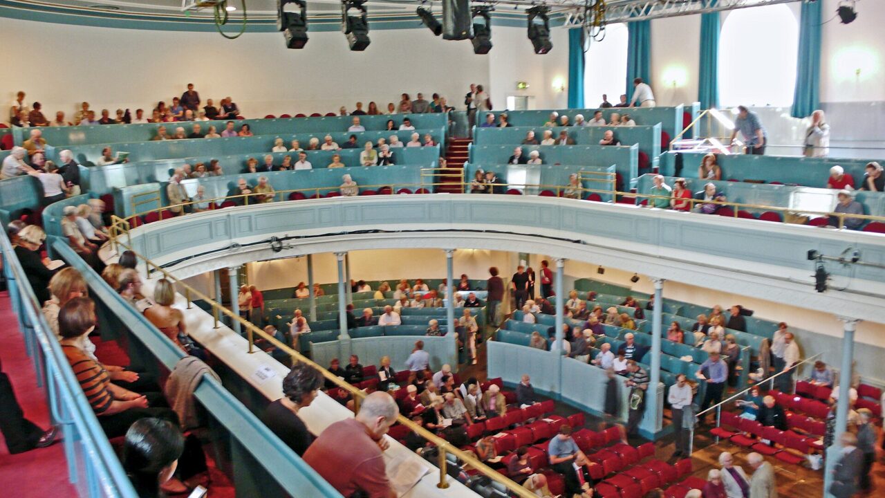 Audience members fill red seats beneath the curved blue balcony inside the Queen's Hall in Edinburgh.