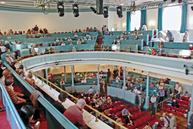 Audience members fill red seats beneath the curved blue balcony inside the Queen's Hall in Edinburgh.