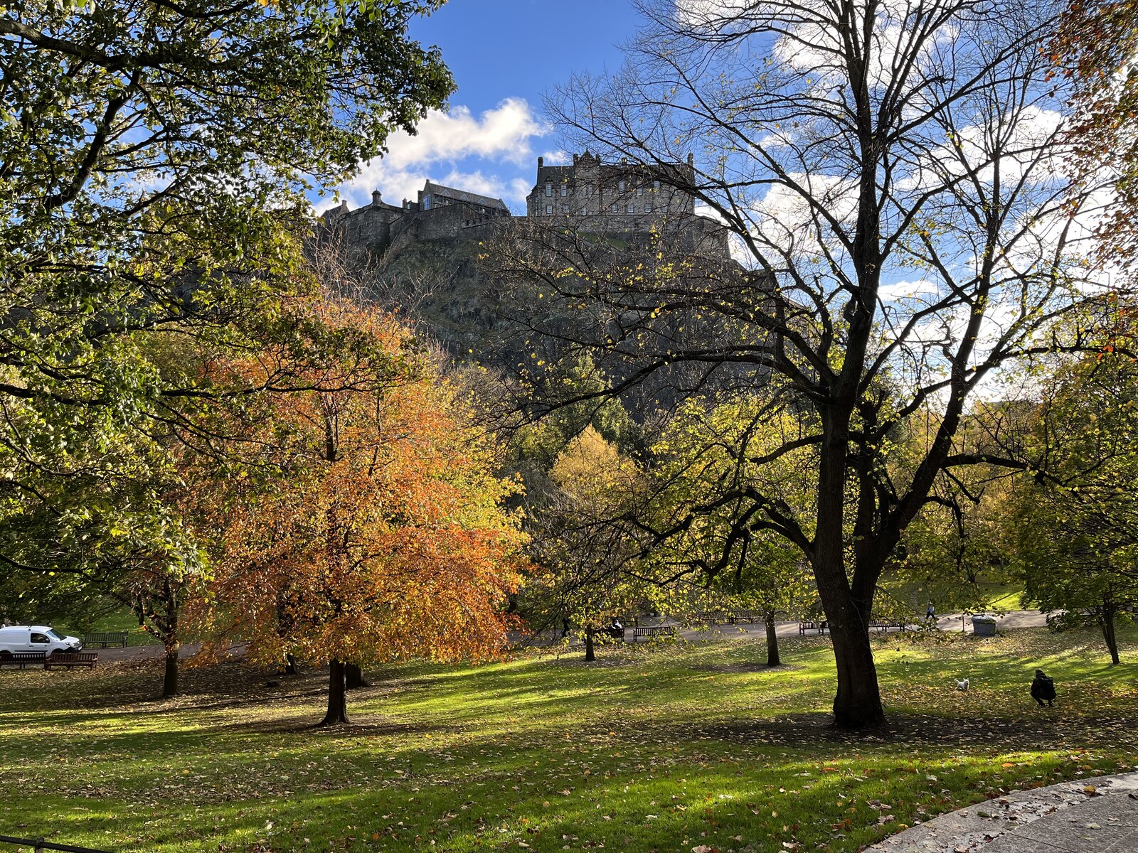Autumn trees turn gold and orange in Princes Street Gardens, with Edinburgh Castle rising above the lawns and paths.