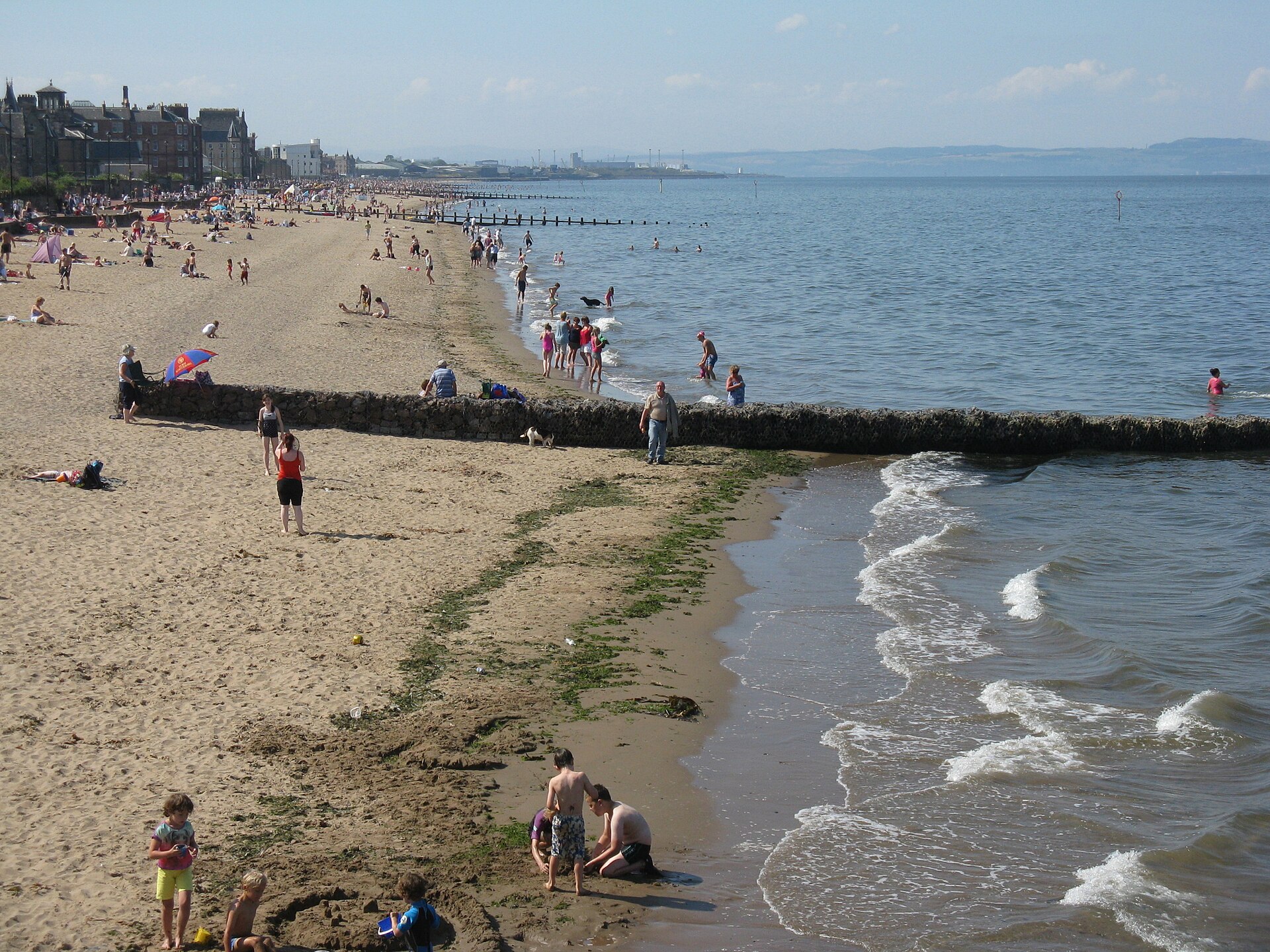 Beachgoers paddle and play in the sand beside a stone groyne on a sunny day at Portobello Beach, Edinburgh.