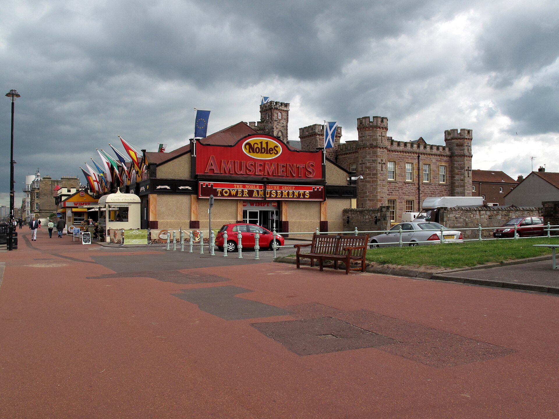 Benches and fluttering flags line the wide paved Portobello promenade beside an amusement arcade and turreted stone building in Edinburgh.