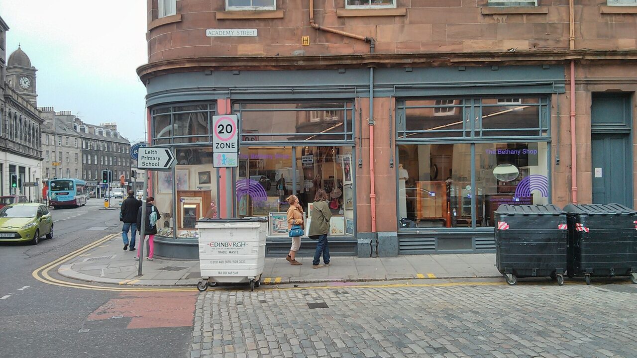 The Bethany charity shop in Edinburgh's Leith has broad corner windows beneath a sandstone tenement, with pedestrians passing outside.