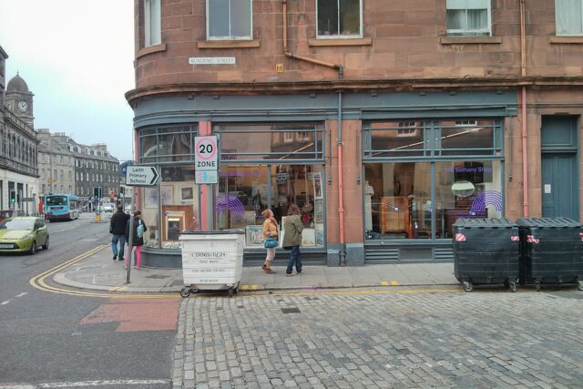 The Bethany charity shop in Edinburgh's Leith has broad corner windows beneath a sandstone tenement, with pedestrians passing outside.