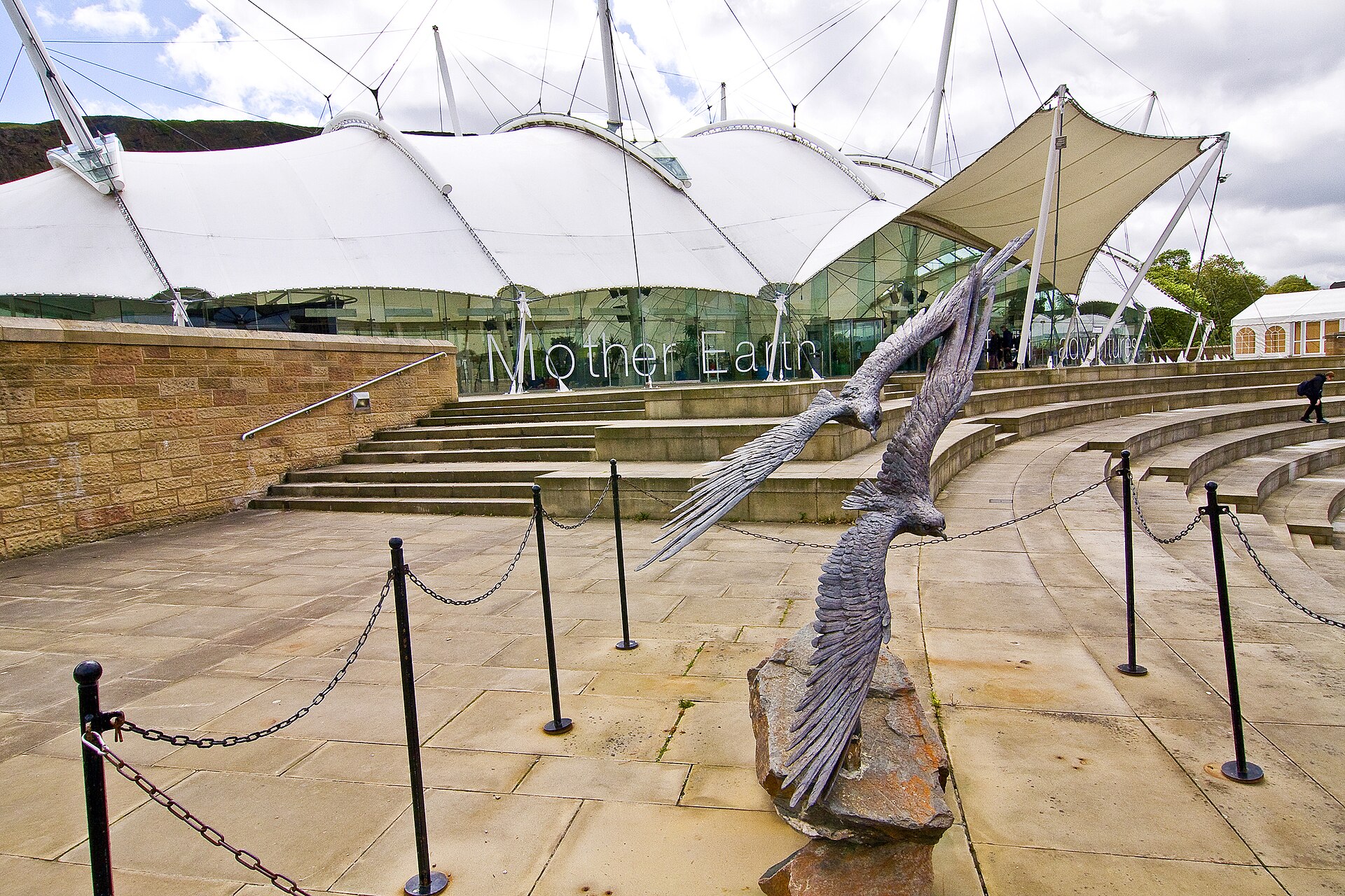 A bird sculpture stands beside curved stone steps outside Dynamic Earth in Edinburgh, beneath its white tented roof.