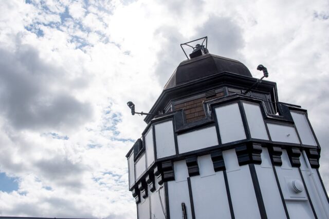 The black-and-white rooftop tower and dark dome of Camera Obscura in Edinburgh rise against a cloudy sky.