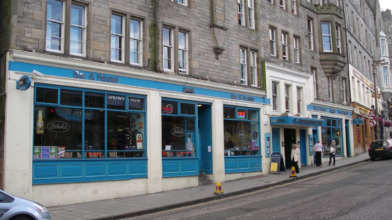 Blue-painted bar and hostel windows line the pavement below Edinburgh stone tenements, with people standing near the entrance.