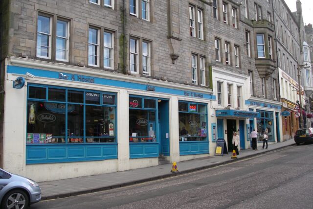 Blue-painted bar and hostel windows line the pavement below Edinburgh stone tenements, with people standing near the entrance.