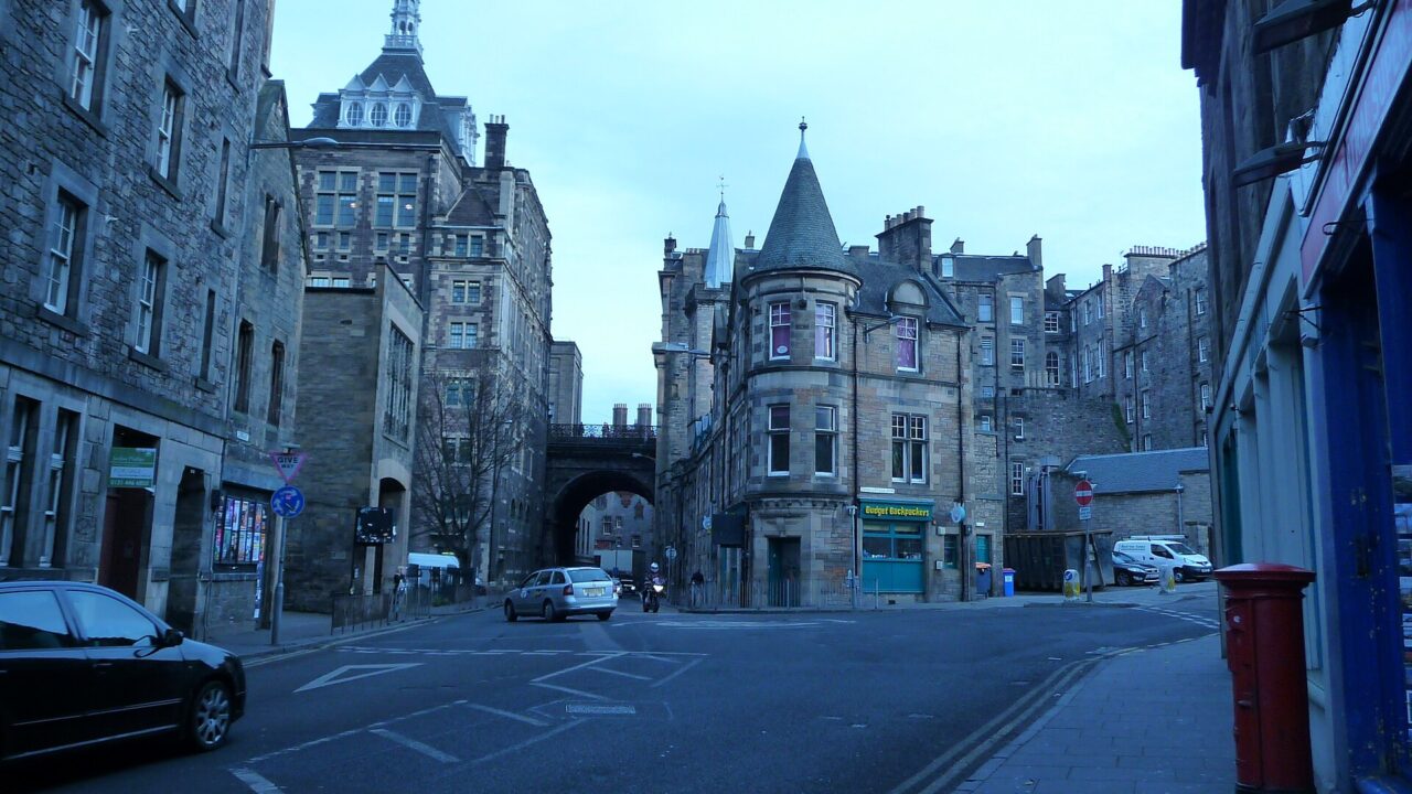 Budget Backpackers occupies a turreted stone corner building beside a bridge in Edinburgh's Old Town, with cars at the junction.