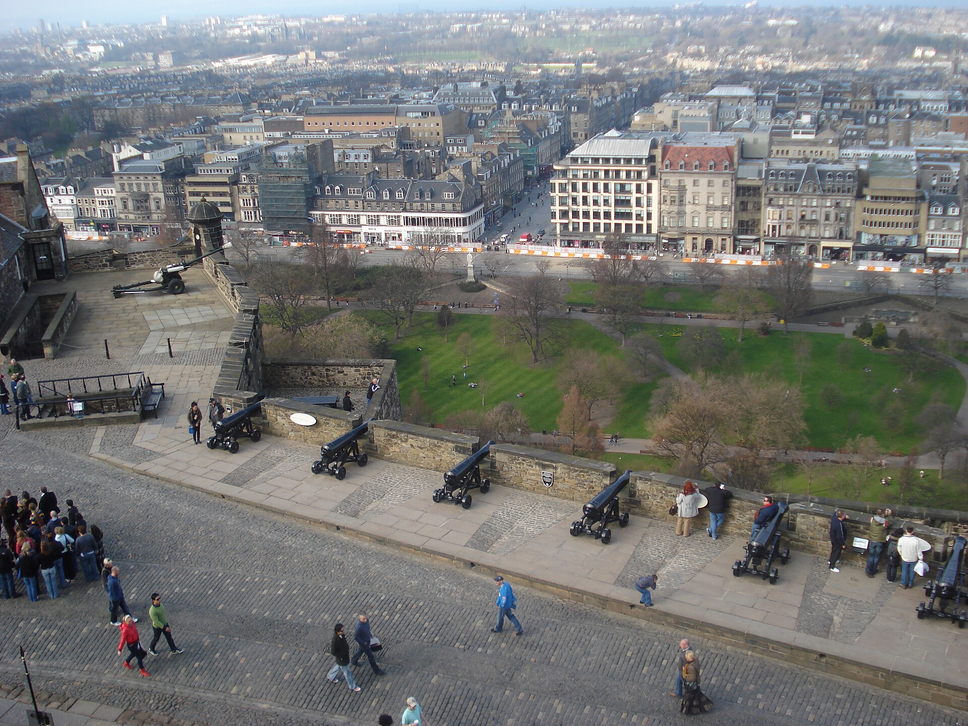Cannons line Edinburgh Castle's ramparts above Princes Street Gardens, with visitors walking beside them and city buildings beyond.