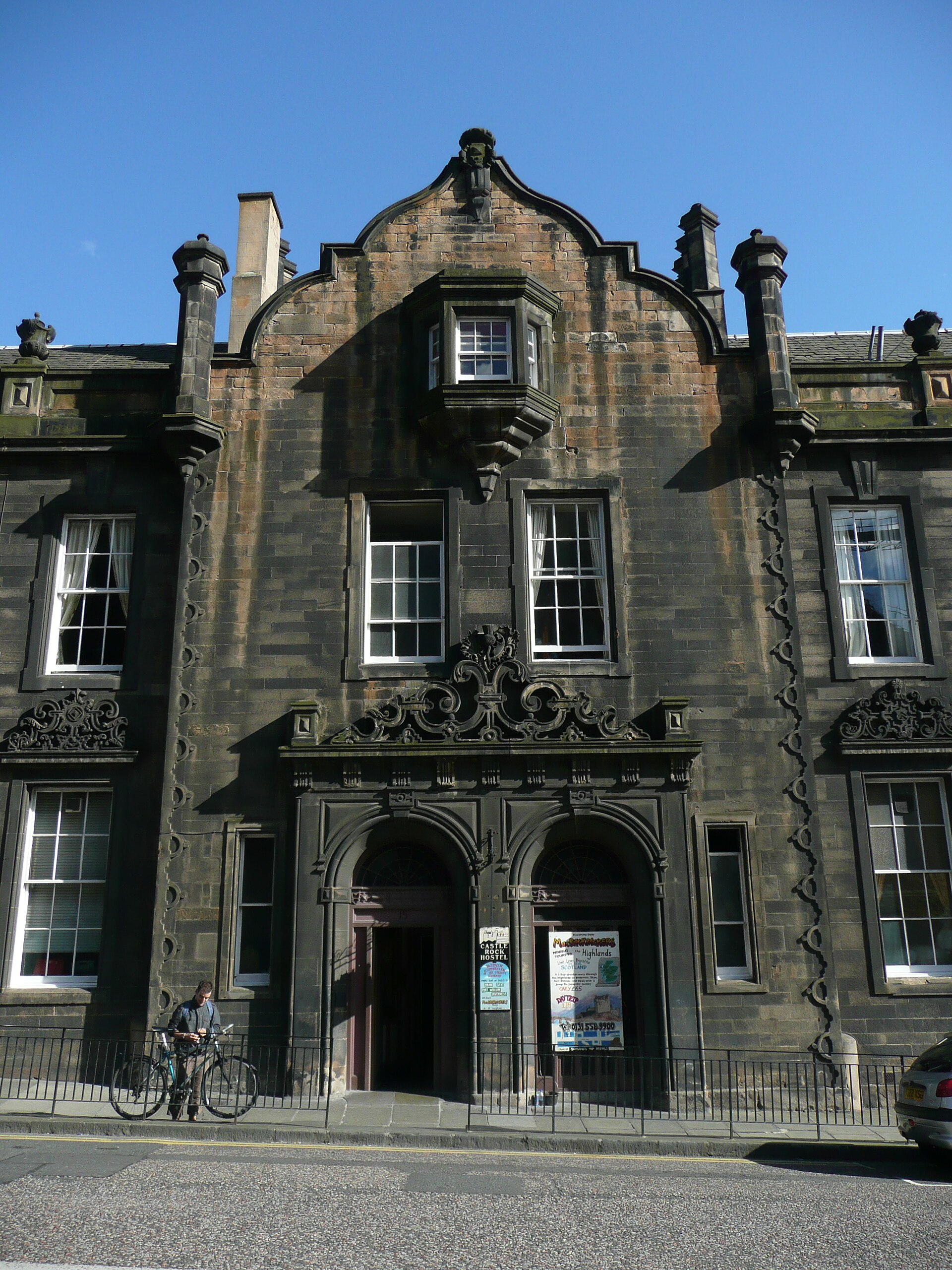 Castle Rock Hostel in Edinburgh has ornate stonework, paired arched doorways and a cyclist standing beside the pavement.