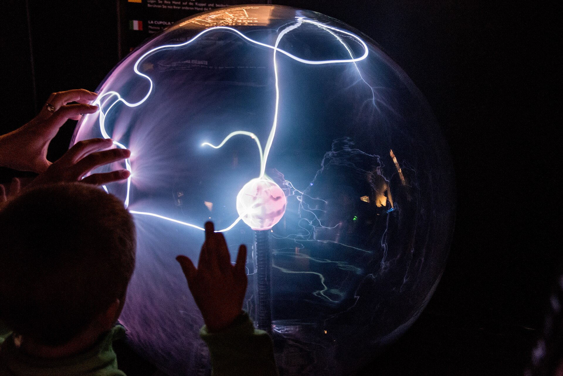 A child touches a glowing plasma globe at Camera Obscura in Edinburgh, with bright filaments branching across the glass.