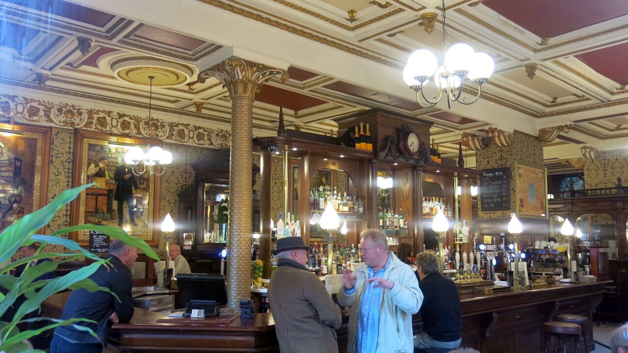 Customers gather at the curved wooden bar of Edinburgh’s Café Royal beneath ornate ceilings, mirrors and globe lamps.
