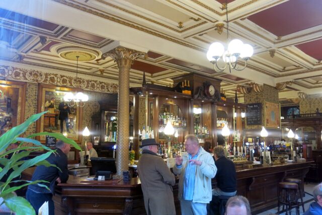 Customers gather at the curved wooden bar of Edinburgh’s Café Royal beneath ornate ceilings, mirrors and globe lamps.