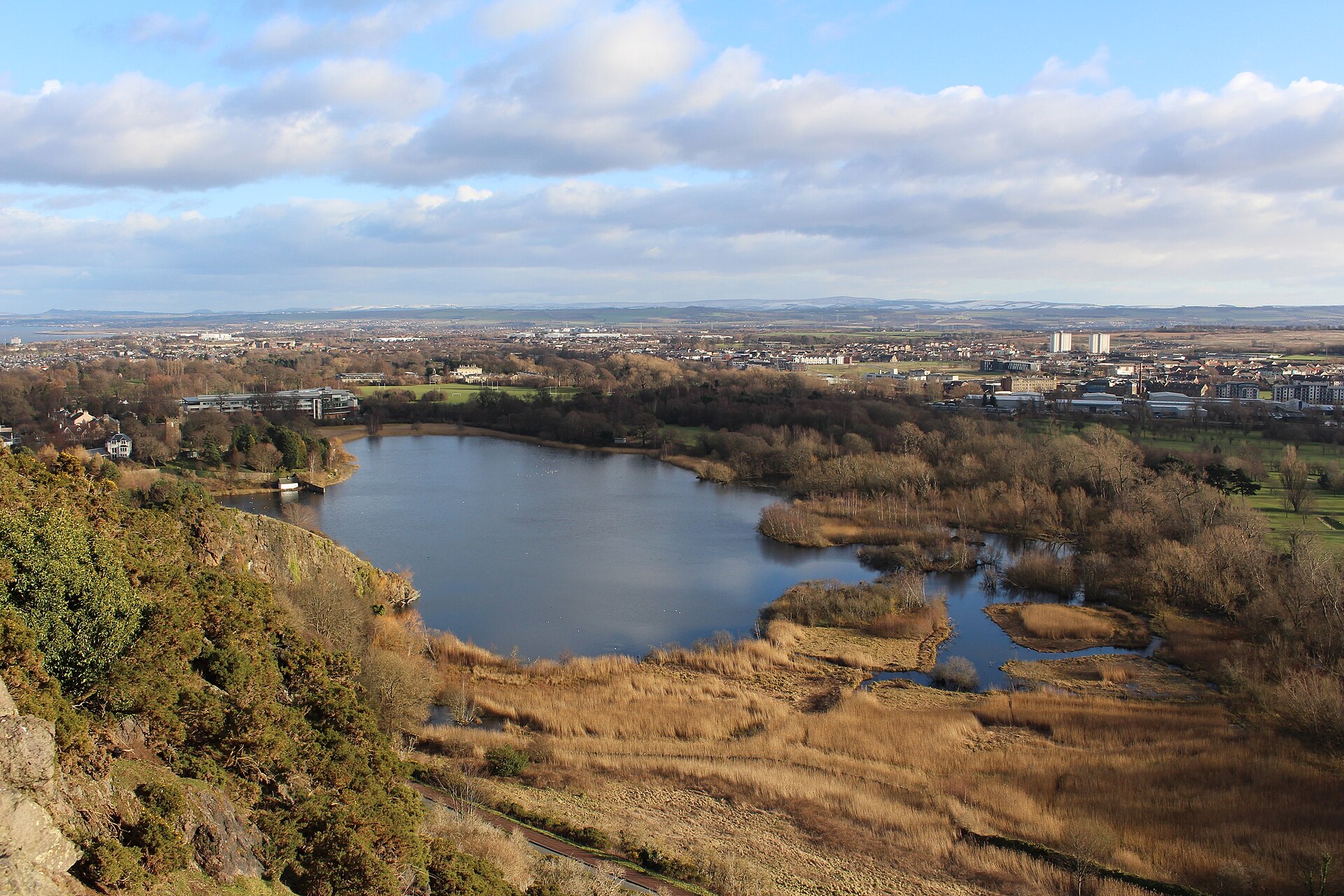 Duddingston Loch lies below a rocky slope, surrounded by golden reedbeds, bare trees and Edinburgh buildings in the distance.