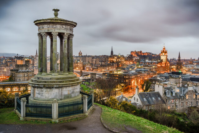 The Dugald Stewart Monument overlooks Edinburgh at dusk, with the illuminated Balmoral clock tower and Edinburgh Castle beyond.