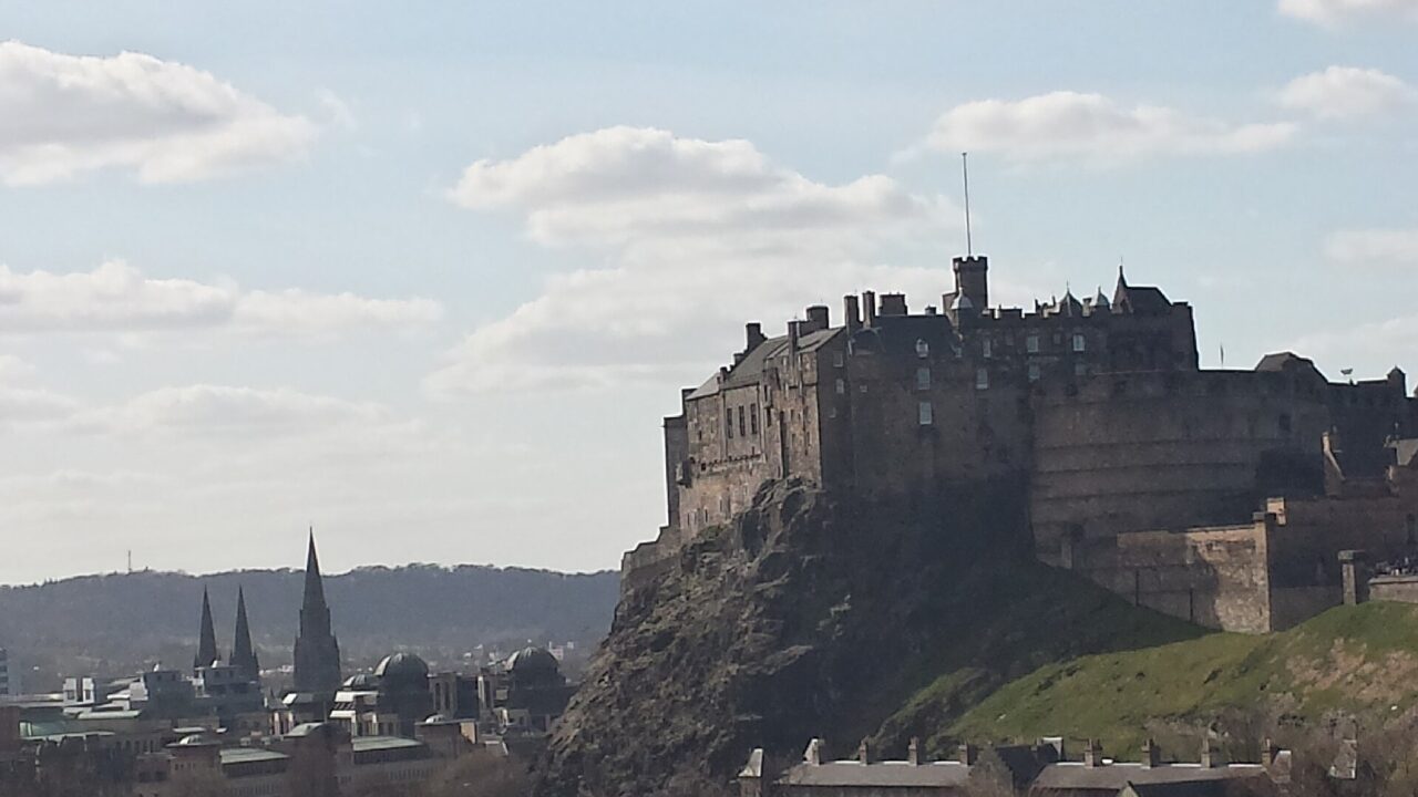 Edinburgh Castle rises above the city's rooftops and church spires, with its rocky slope visible beneath a partly cloudy sky.