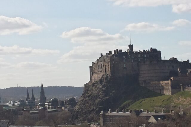 Edinburgh Castle rises above the city's rooftops and church spires, with its rocky slope visible beneath a partly cloudy sky.