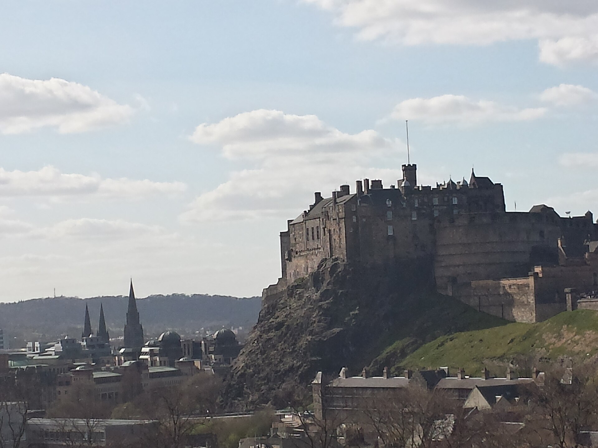 Edinburgh Castle rises above the city's rooftops and church spires, with its rocky slope visible beneath a partly cloudy sky.