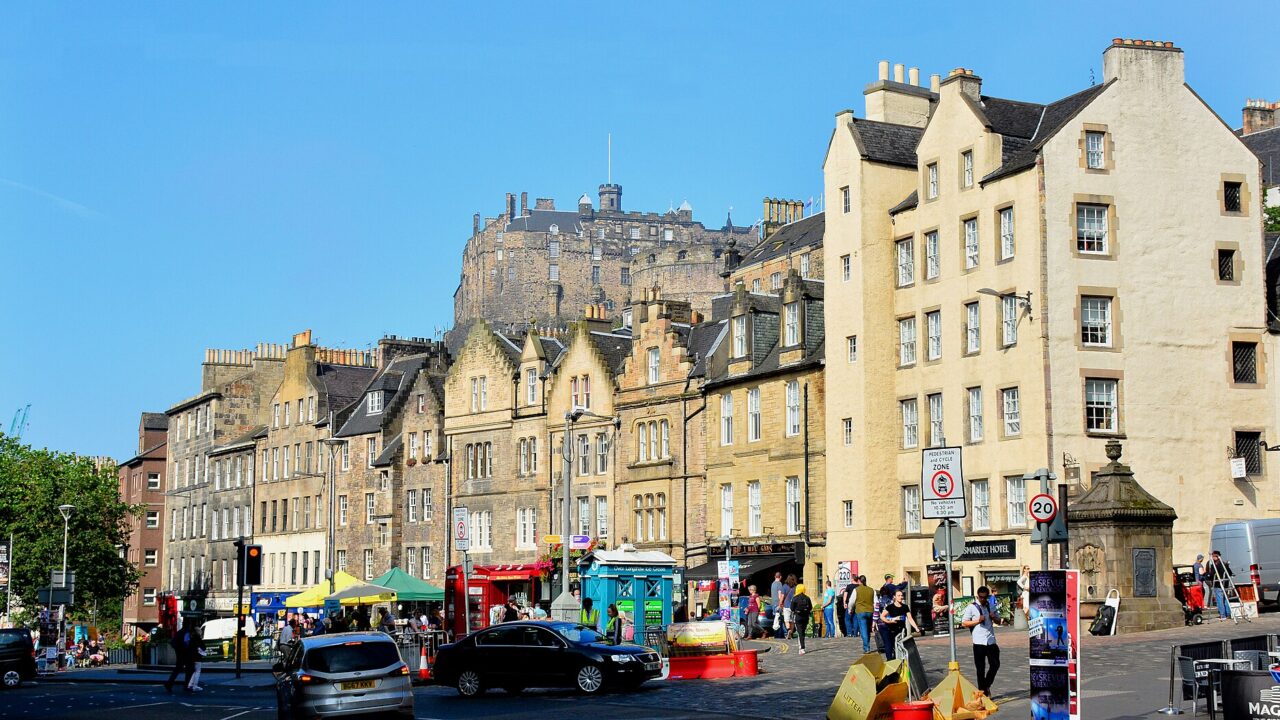 Edinburgh Castle rises above gabled Grassmarket buildings, colourful shopfronts and pedestrians beneath a clear blue sky.