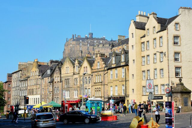 Edinburgh Castle rises above gabled Grassmarket buildings, colourful shopfronts and pedestrians beneath a clear blue sky.