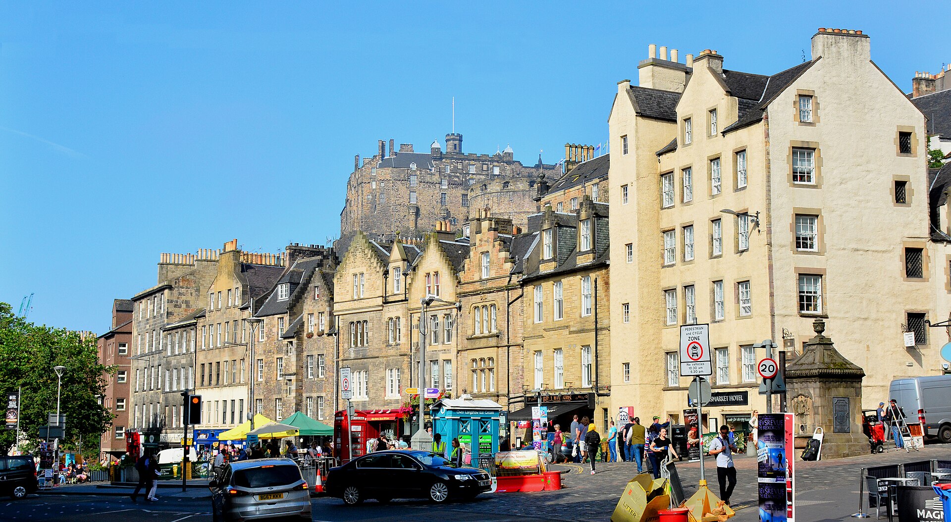 Edinburgh Castle rises above gabled Grassmarket buildings, colourful shopfronts and pedestrians beneath a clear blue sky.