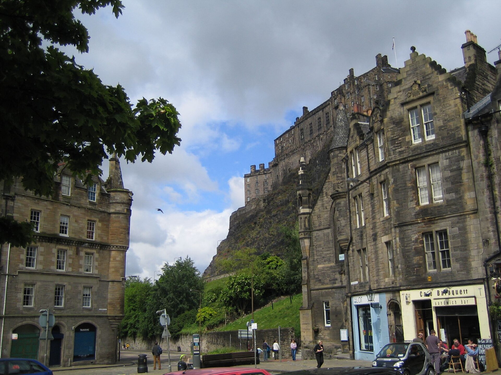 Edinburgh Castle towers above stone buildings and pedestrians at the foot of the rock beside the Grassmarket.