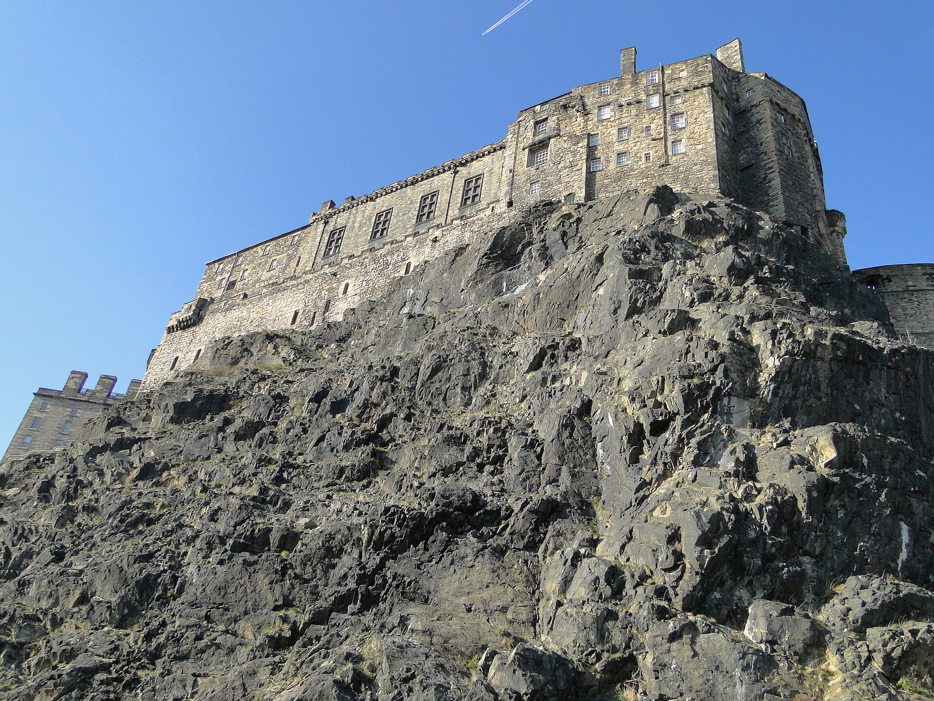 Edinburgh Castle's stone buildings stand directly above jagged exposed rock in a low-angle view beneath a clear blue sky.