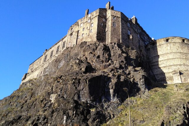Edinburgh Castle's sunlit stone buildings and rounded bastion rise above the rugged face of Castle Rock under blue sky.