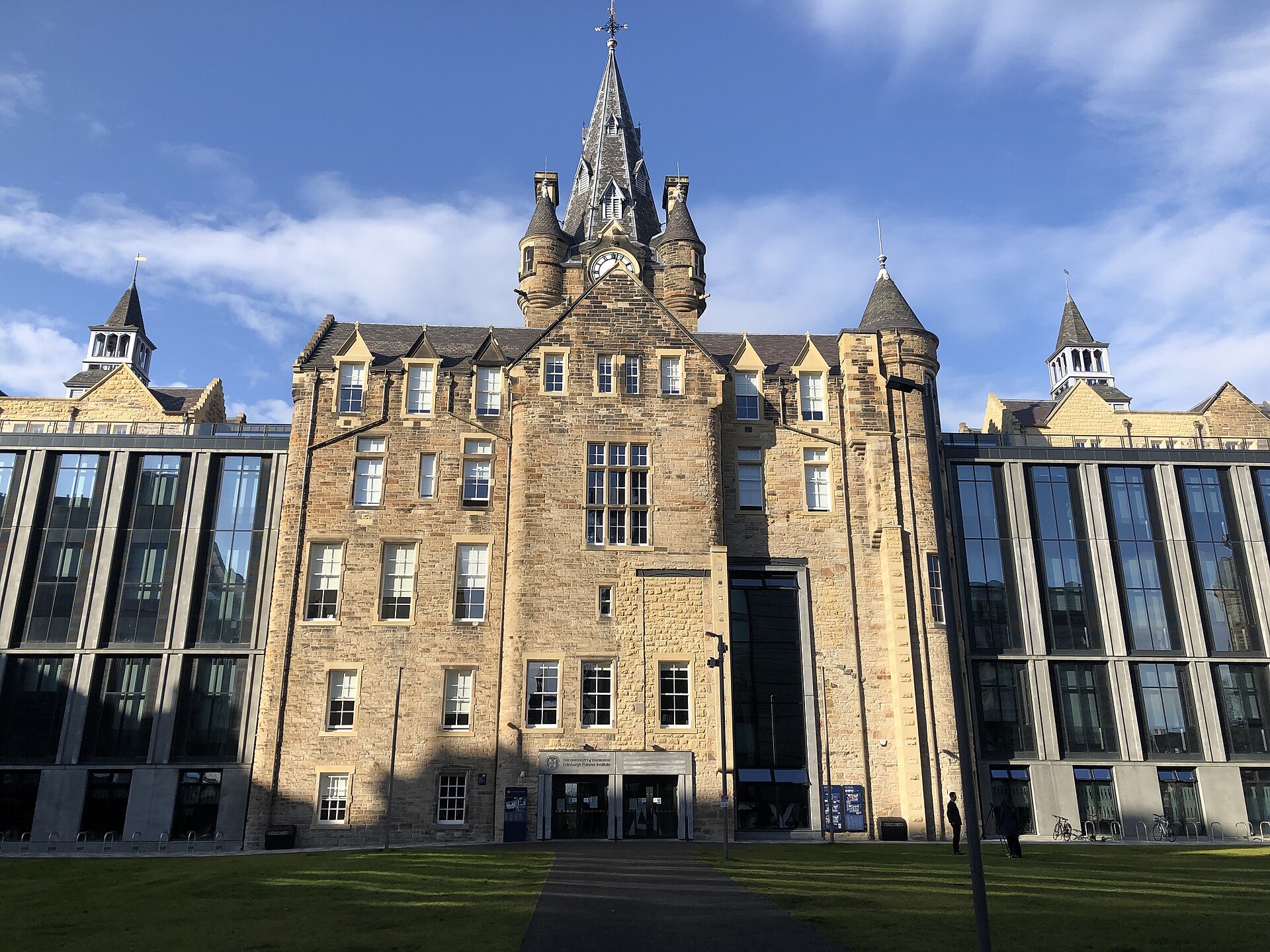 Edinburgh Futures Institute's stone clock tower rises above its entrance, flanked by modern glass wings beside a green lawn.