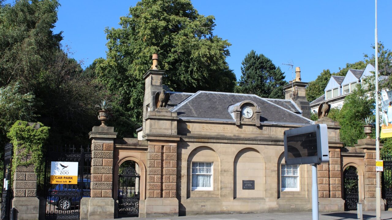 Edinburgh Zoo gate lodge has a central clock, stone arches and iron gates, with leafy trees rising behind.