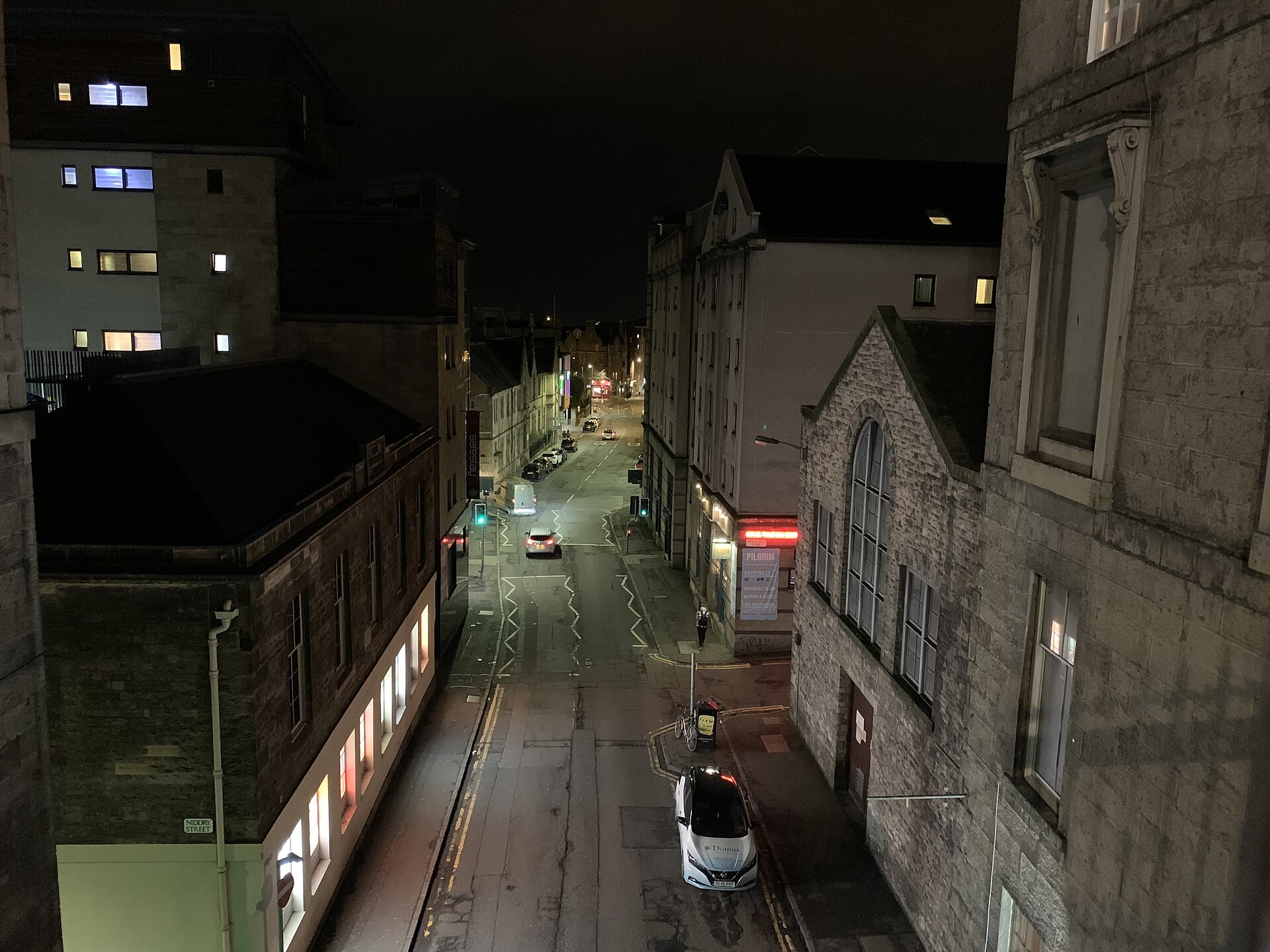 Edinburgh's Cowgate stretches between stone buildings at night, with lit windows, parked cars and pedestrians beneath streetlights.