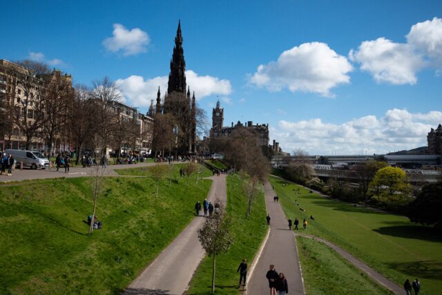 Edinburgh's Scott Monument rises beside Princes Street Gardens, where visitors walk along paths between sloping lawns beneath scattered clouds.