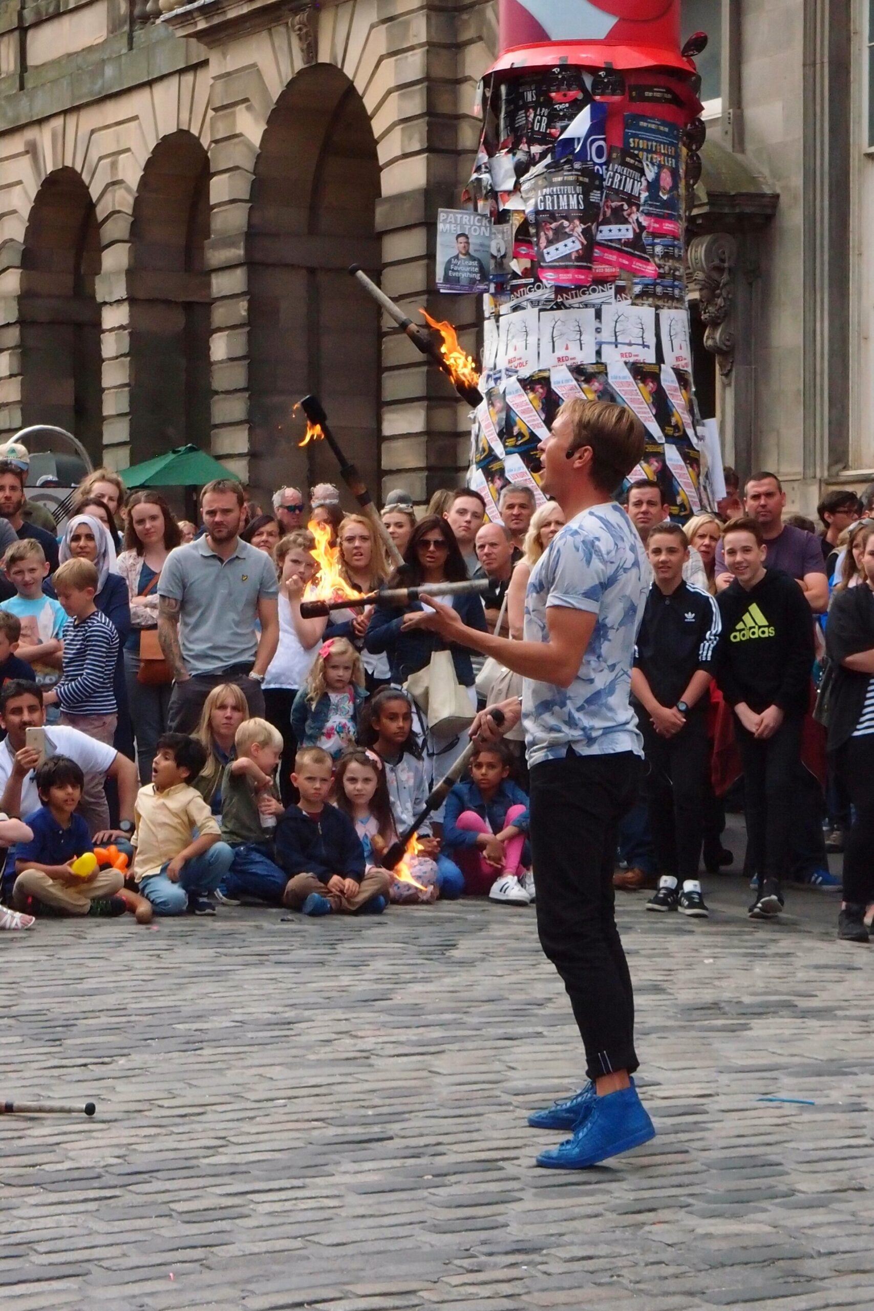 A fire juggler entertains a crowd on Edinburgh's Royal Mile beside a column covered in Fringe show posters.
