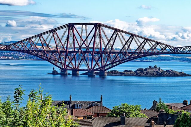 The Forth Bridge spans blue water above Inchgarvie, seen over rooftops and trees in South Queensferry.