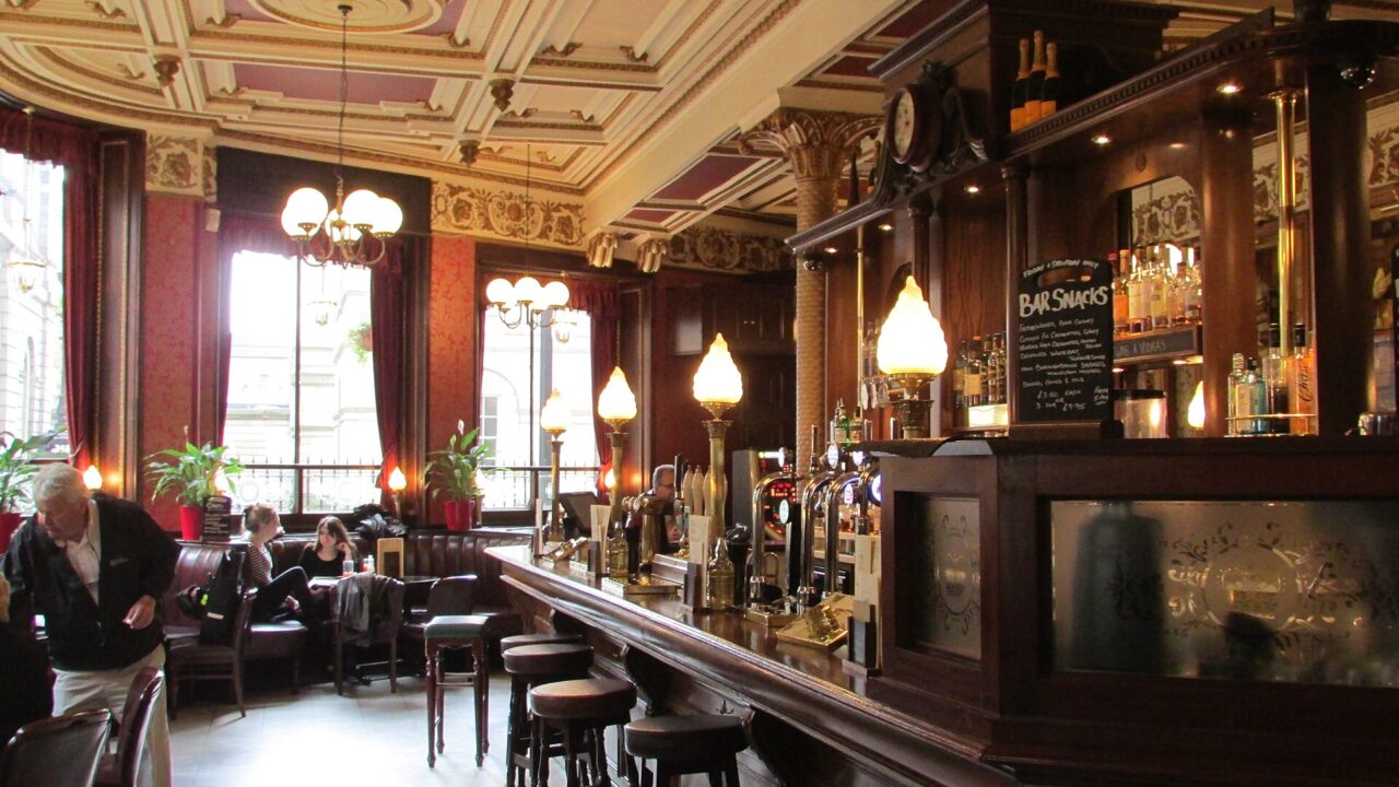 Glowing lamps line the polished wooden counter inside Edinburgh's Café Royal, with stools and customers beside tall windows.