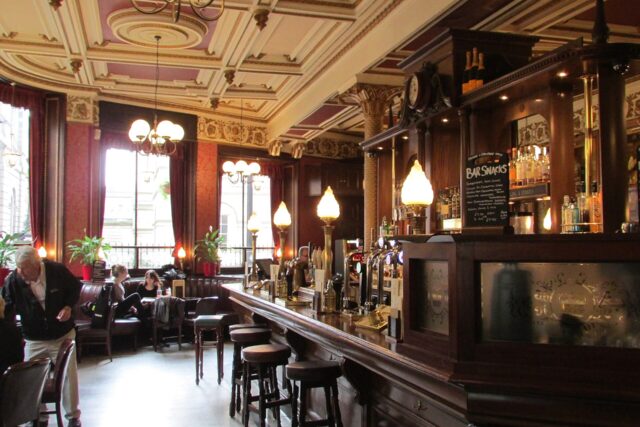 Glowing lamps line the polished wooden counter inside Edinburgh's Café Royal, with stools and customers beside tall windows.