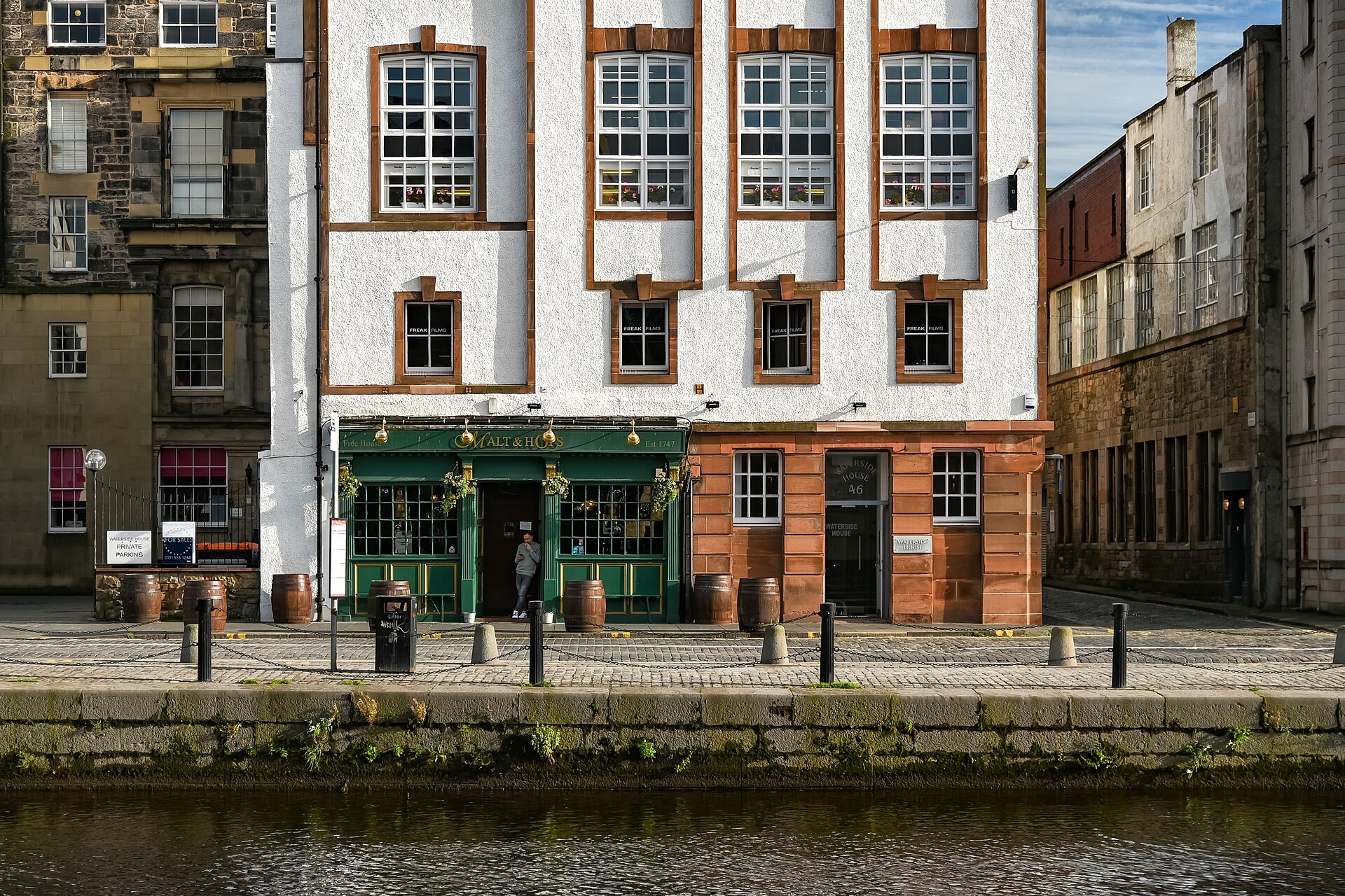 The green Malt and Hops pub frontage and barrel tables face the Water of Leith beneath a tall white building.