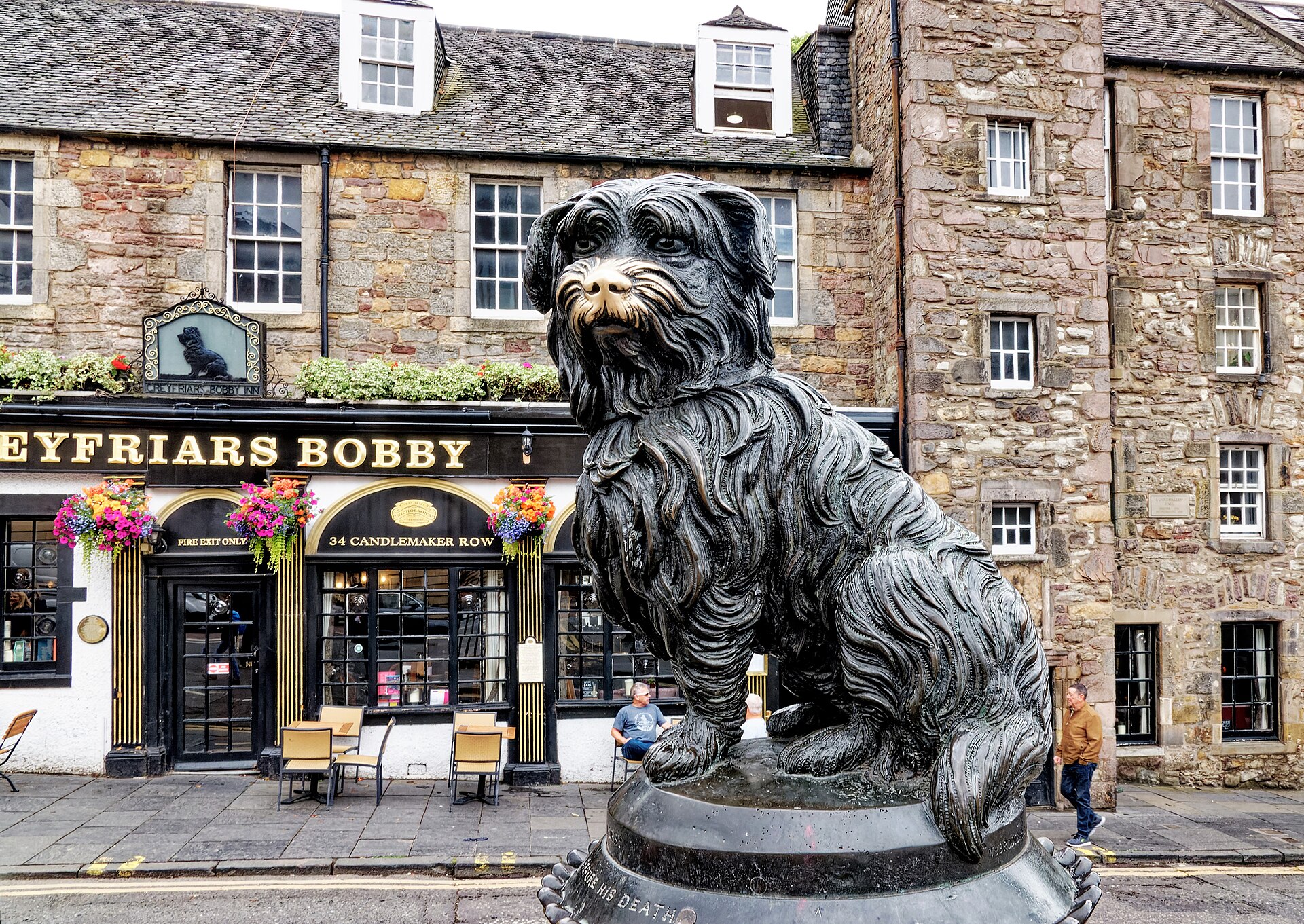 Greyfriars Bobby's bronze statue on Candlemaker Row in Edinburgh, with stone buildings and the pub behind it.