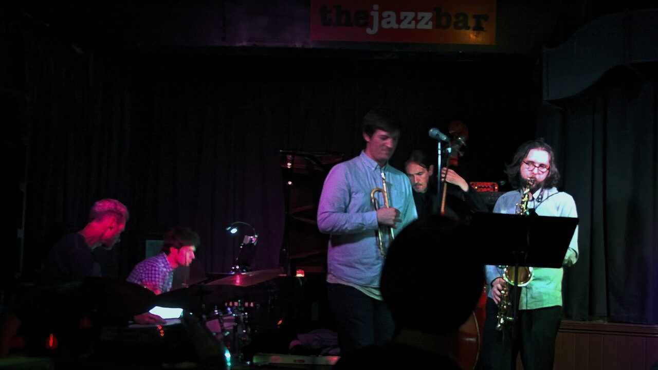 A jazz band with drums, piano, trumpet, double bass and saxophone plays on the small stage of The Jazz Bar in Edinburgh