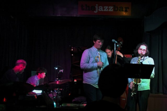 A jazz band with drums, piano, trumpet, double bass and saxophone plays on the small stage of The Jazz Bar in Edinburgh