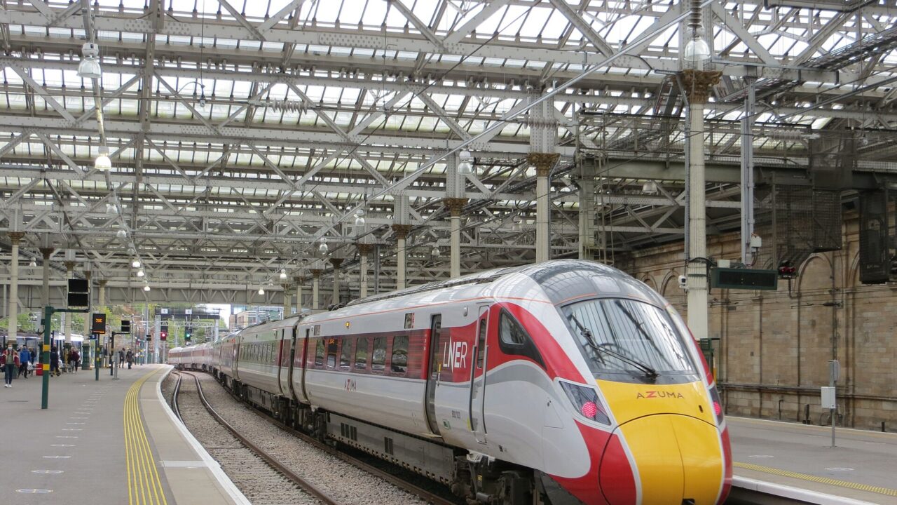 An LNER Azuma train stands beside a curving platform under the glass roof at Edinburgh Waverley.