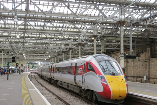 An LNER Azuma train stands beside a curving platform under the glass roof at Edinburgh Waverley.