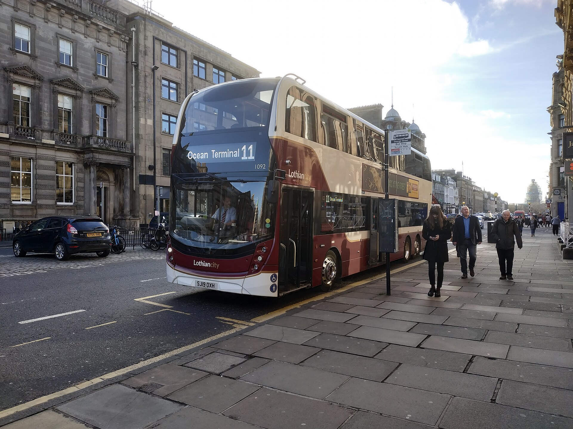 A madder-and-white Lothian double-decker waits at a central Edinburgh bus stop beside pedestrians and stone buildings.