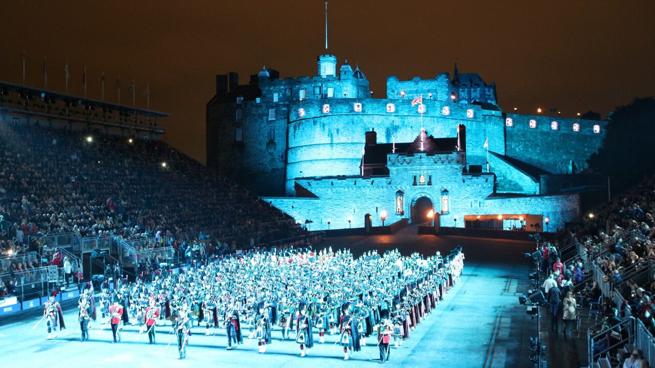 Massed pipers and drummers fill Edinburgh Castle Esplanade between spectator stands beneath the castle walls illuminated in blue.