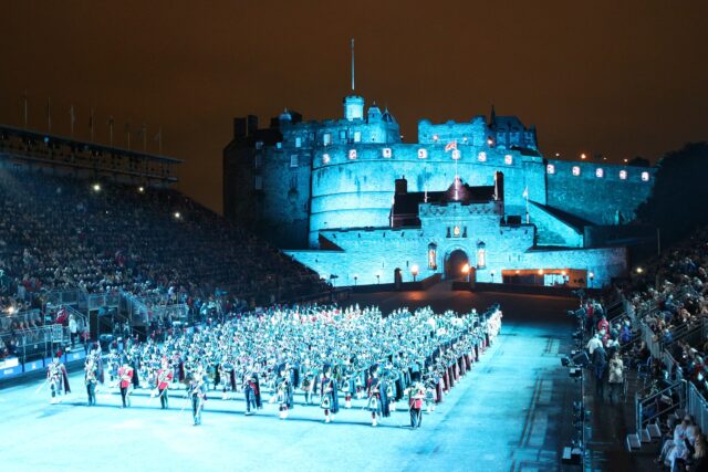 Massed pipers and drummers fill Edinburgh Castle Esplanade between spectator stands beneath the castle walls illuminated in blue.