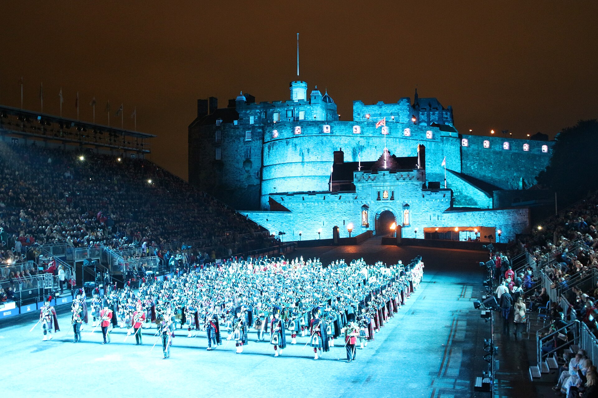 Massed pipers and drummers fill Edinburgh Castle Esplanade between spectator stands beneath the castle walls illuminated in blue.