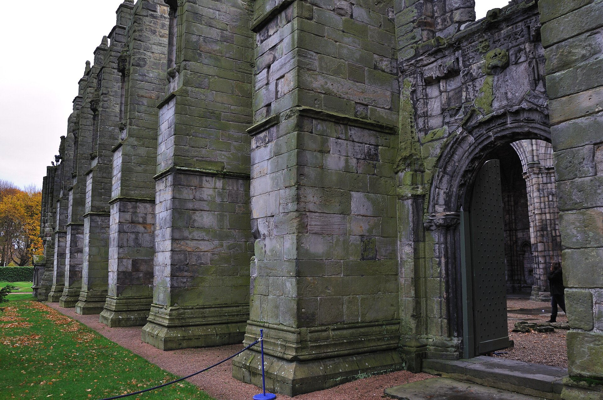 Massive stone buttresses and an arched doorway along Holyrood Abbey's surviving wall in Edinburgh, with autumn leaves outside.