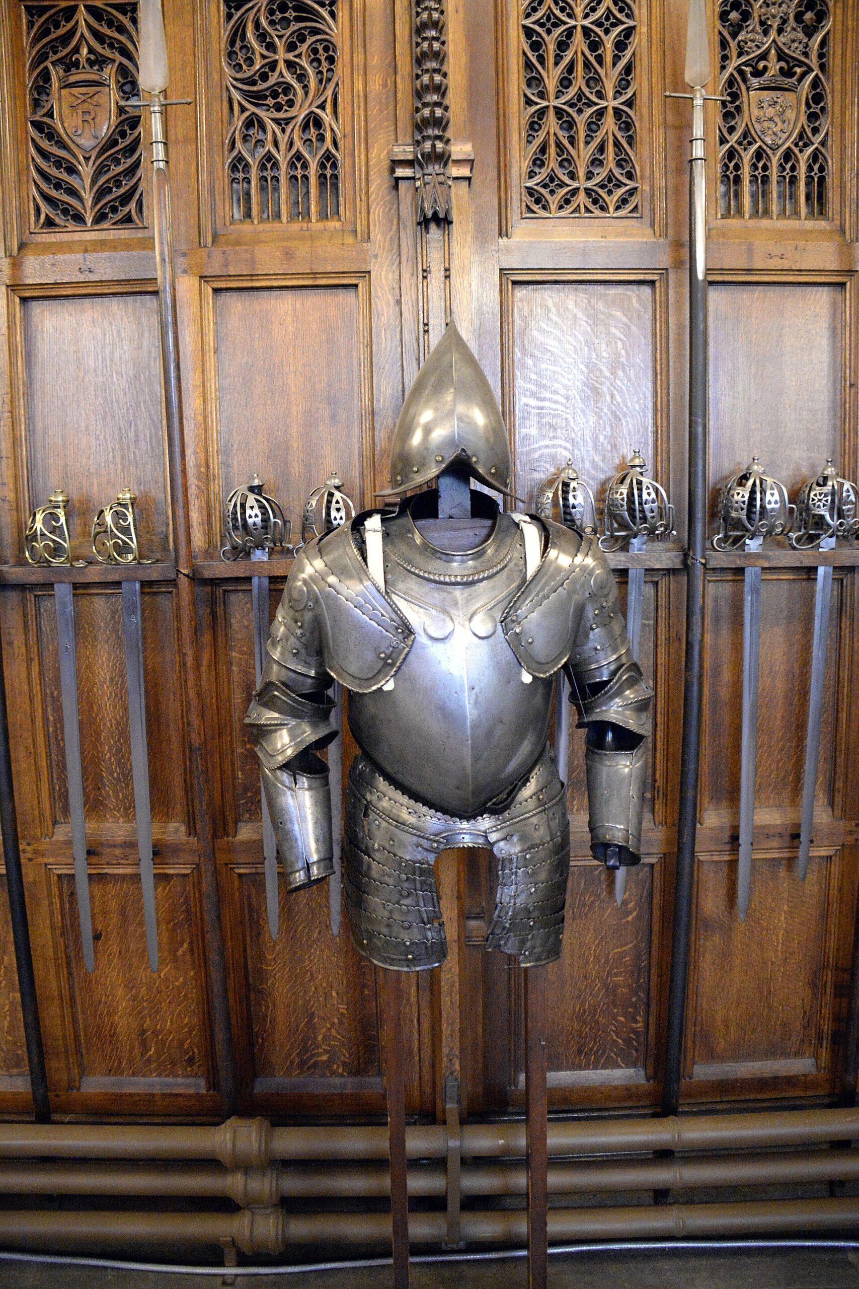 Metal armour and a pointed helmet stand among swords and polearms against carved panelling in Edinburgh Castle's Great Hall.